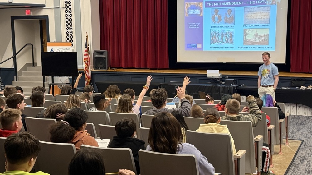 students in an auditorium with hands raised for a standing presenter