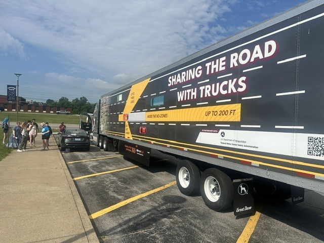 an image with a semi truck with the words on the side "Sharing the Road with Trucks." The semi is in the parking lot of Parkersburg South High School. Some students can be seen at a distance viewing the truck.