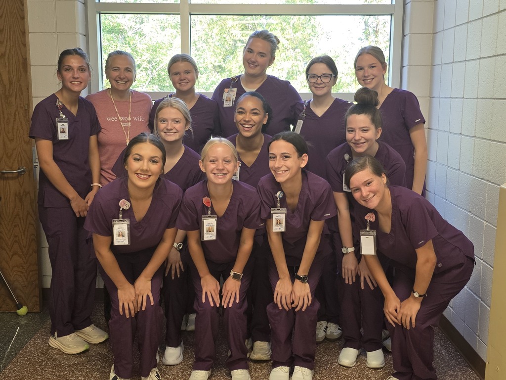 Group of students are wearing matching purple medical scrubs pointing.