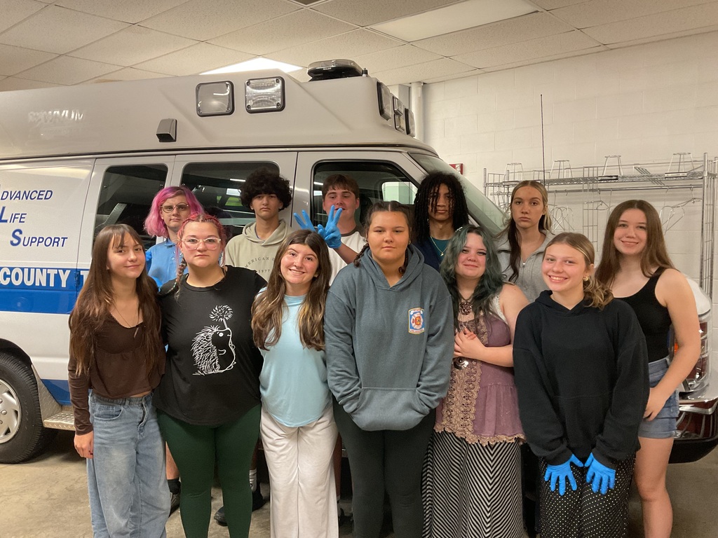 A group of approximately 12 students pose for a photo inside an EMS training area beside a large white ambulance.