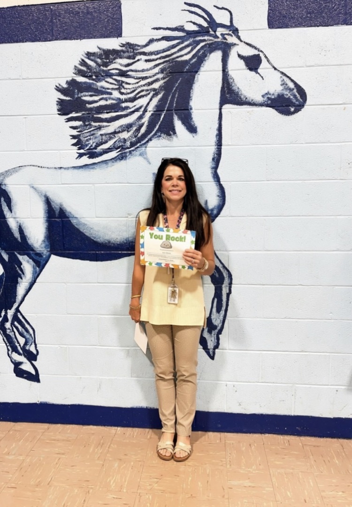 Teacher standing in front of a mural of the Madison Mustang holding a “You Rock” certificate.