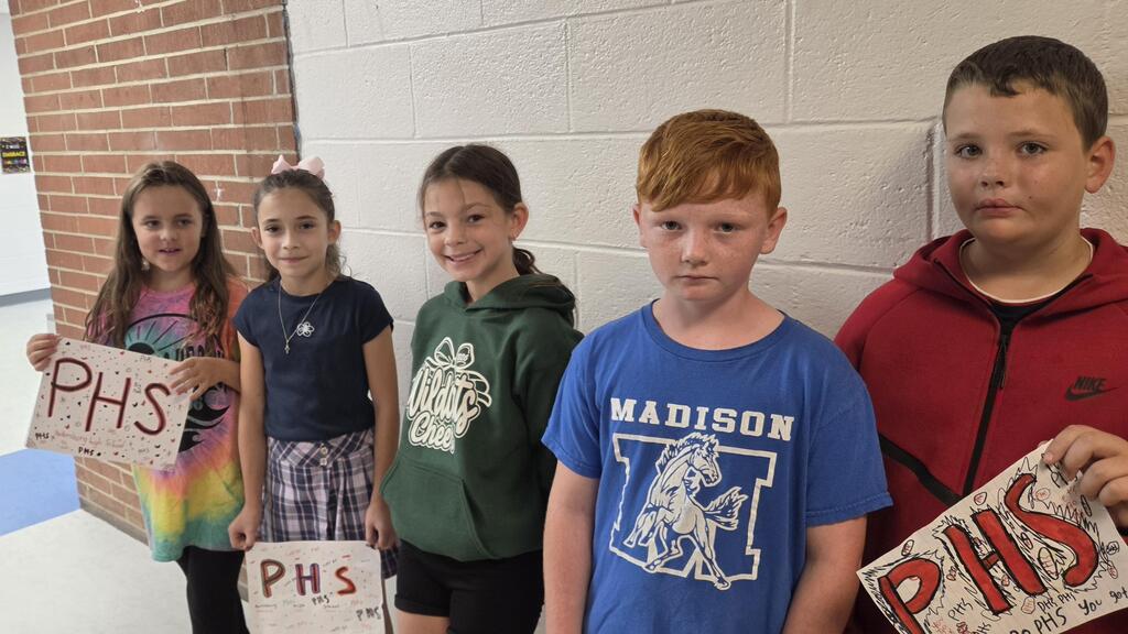 Five students smiling and holding signs before the band walks down the hall.