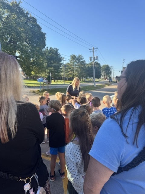 woman speaking to school children outside with large green trees in the background