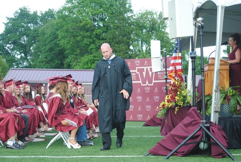 teacher, in cap and gown, leading students to line up to receive diplomas