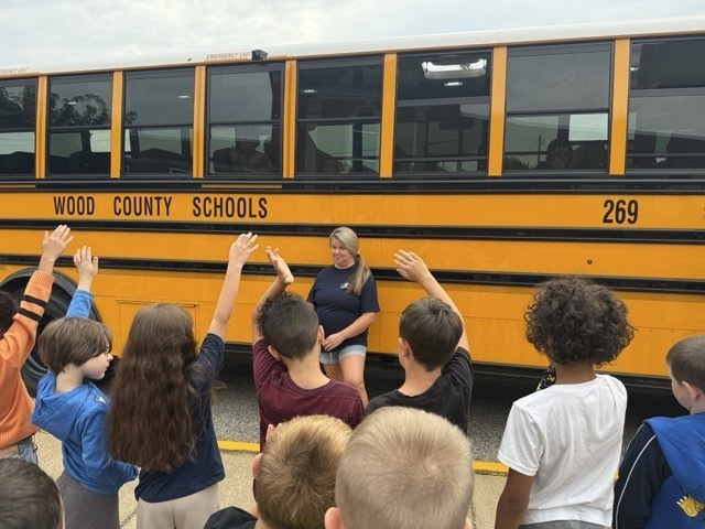 students, with raised hands for questions, standing near a yellow school bus