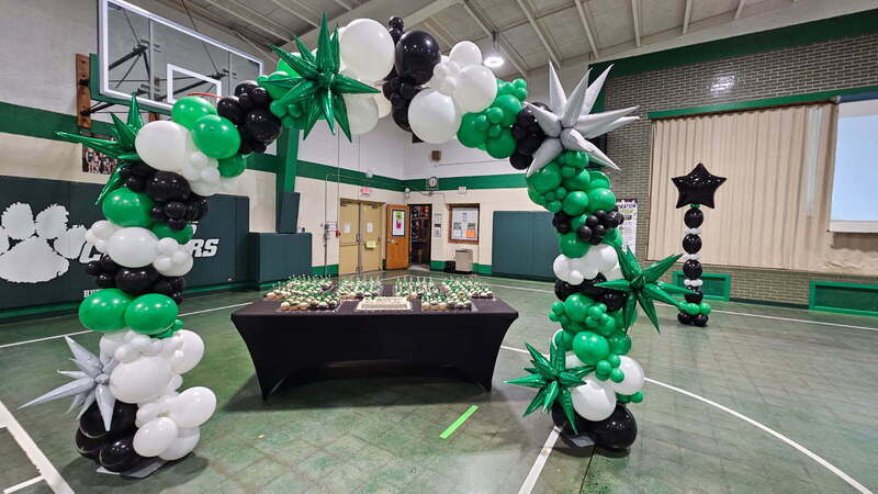 balloon arch in white, black, and green colors hovering over a table with a sheet cake