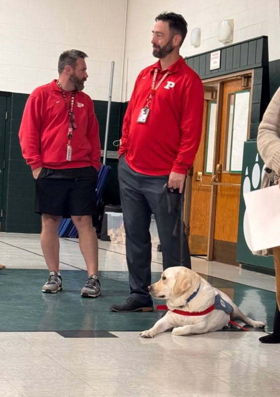 two men standing with a therapy dog which is laying on a gymnasium floor