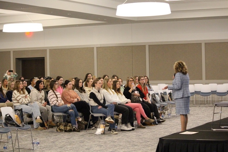 Superintendent Wood County Schools addresses a group of senior women. Superintendent is standing while students are seated facing the stage.