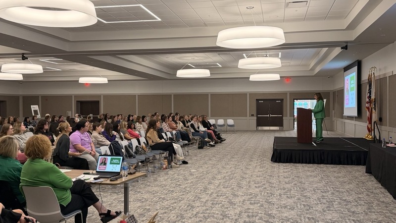 Woman standing on platform in front of podium, speaking to seated students