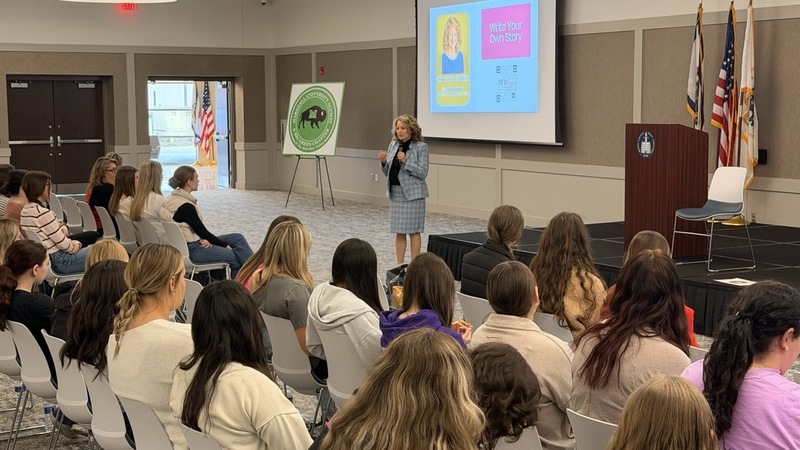 Woman addressing seated students. Woman in blue professional attire, holding microphone with a projector screen behind her.