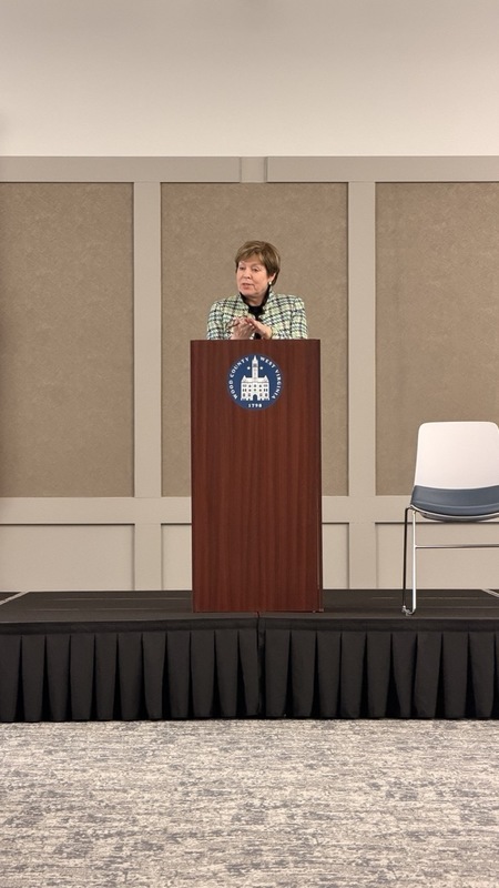 Woman standing on small platform behind Lecturn speaking to a group of student students