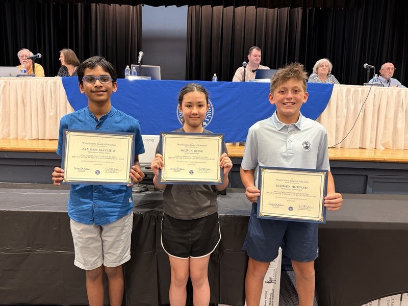 Three students holding certificate with Board of Education sitting in background