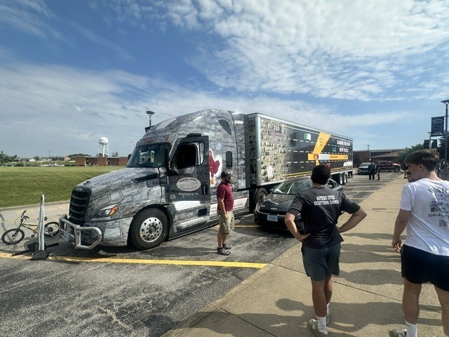 image of a semi truck parked in a high school parking lot with blue skies and light clouds above. The school campus off in the distance.