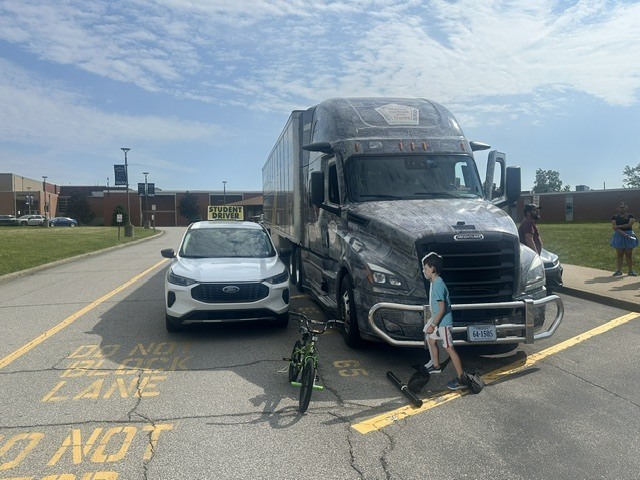 image of a semi truck parked in a high school parking lot with blue skies and light clouds above. The school campus off in the distance.