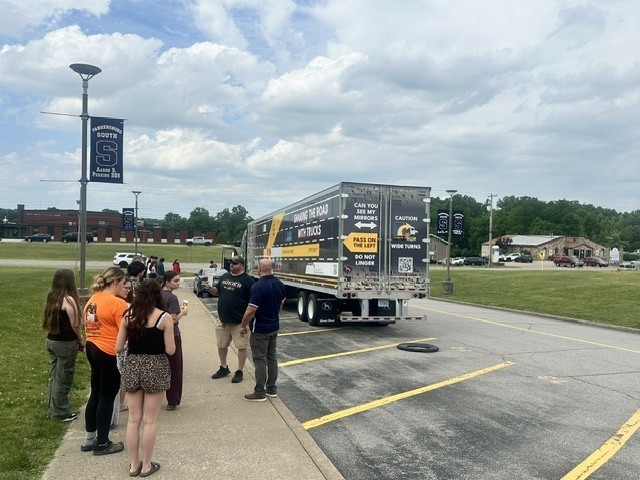 image of a semi truck parked in a high school parking lot with blue skies and light clouds above. The school campus off in the distance.