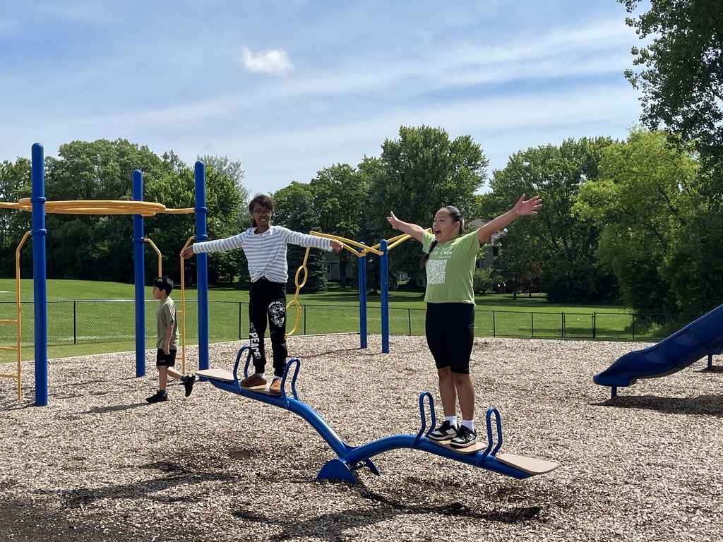 two students stand on a see saw on an outdoor playground with arms spread wide