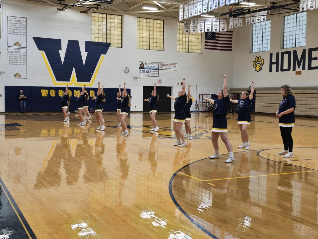 Cheerleaders stand in the middle of the gym floor, facing the students, performing the fight song dance.