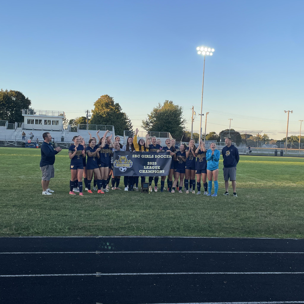 Girls Soccer Team holding a blue banner with the Wildcat logo (holding Woodmore and a soccer ball) on the left with "SBC Girls Soccer 2025 League Champion" written on it. The team has very exceited faces and are clearly cheering. The picture was taken on the main field at Woodmore.
