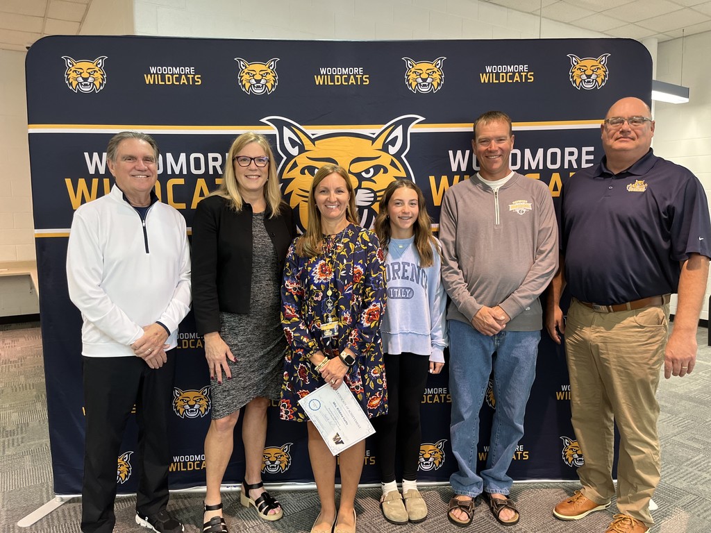 From left to right: Superintendent Mr. Mock, High School Principal Mrs. Coil, Mrs. Curtis and family, Board President Mr. Miller. They're all standing in front of the Woodmore Wildcats backdrop in the high school media center.