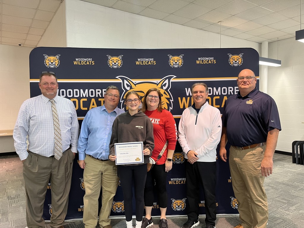 From left to right: Middle School Principal Mr. Barrett, Evelynn and family, Superintendent Mr. Mock, Board President Mr. Miller. They're all standing in front of the Woodmore Wildcats backdrop in the high school media center.