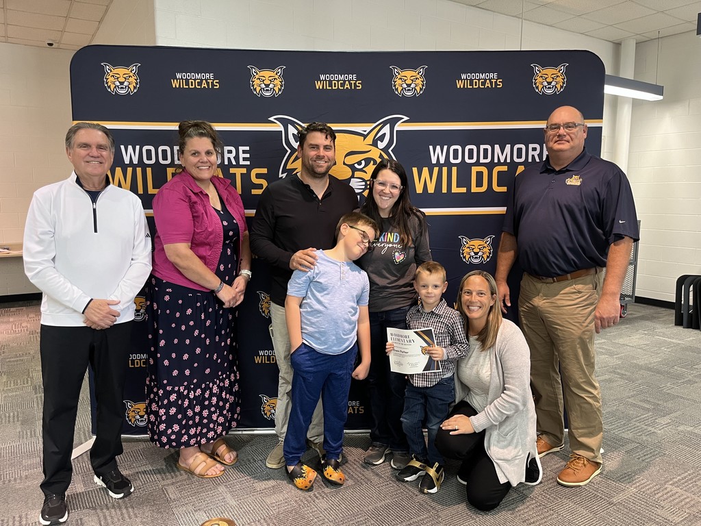 From left to right: Superintendent Mr. Mock, Elementary Principal Mrs. Lafontaine, Owen and family, teacher Mrs. Clouse, Board President Mr. Miller. They're all standing in front of the Woodmore Wildcats backdrop in the high school media center.