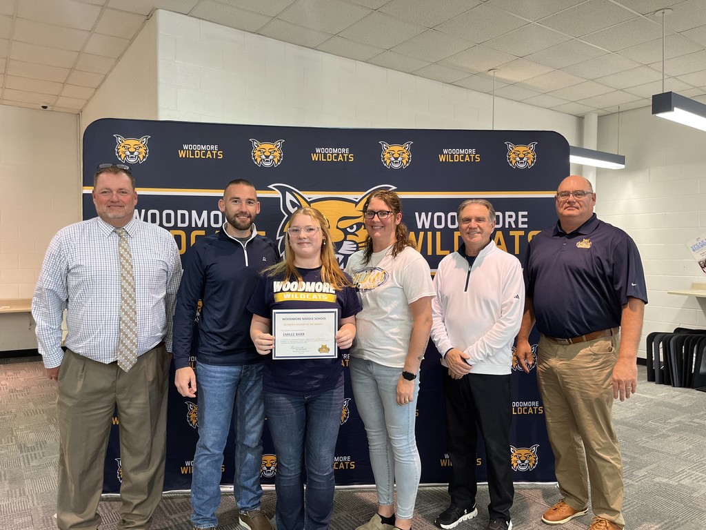 From left to right: Middle School Principal Mr. Barrett, Emilee and family, Superintendent Mr. Mock, Board President Mr. Miller. They're all standing in front of the Woodmore Wildcats backdrop in the high school media center.