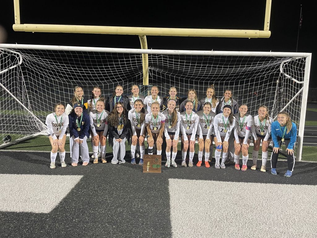 Picture of the girls soccer team in front of the net with their Regional Champs trophy on the ground in front of them . They're all also wearing medals.
