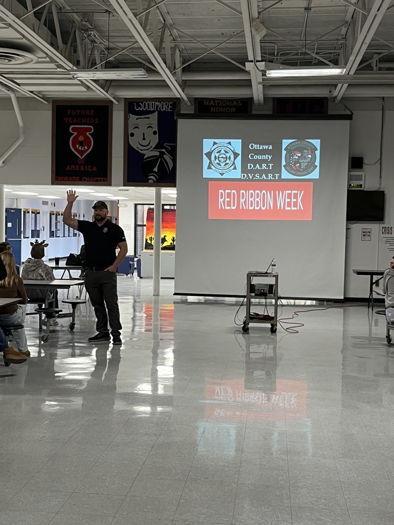 Deputy Trevor Johnson speaks to students in the high school cafeteria. There is a screen behind him with logos for Ottawa County D.A.R.T. and D.V.S.A.R.T. along with a red rectangle with the words "Red Ribbon Week" in white in the middle of the rectangle.