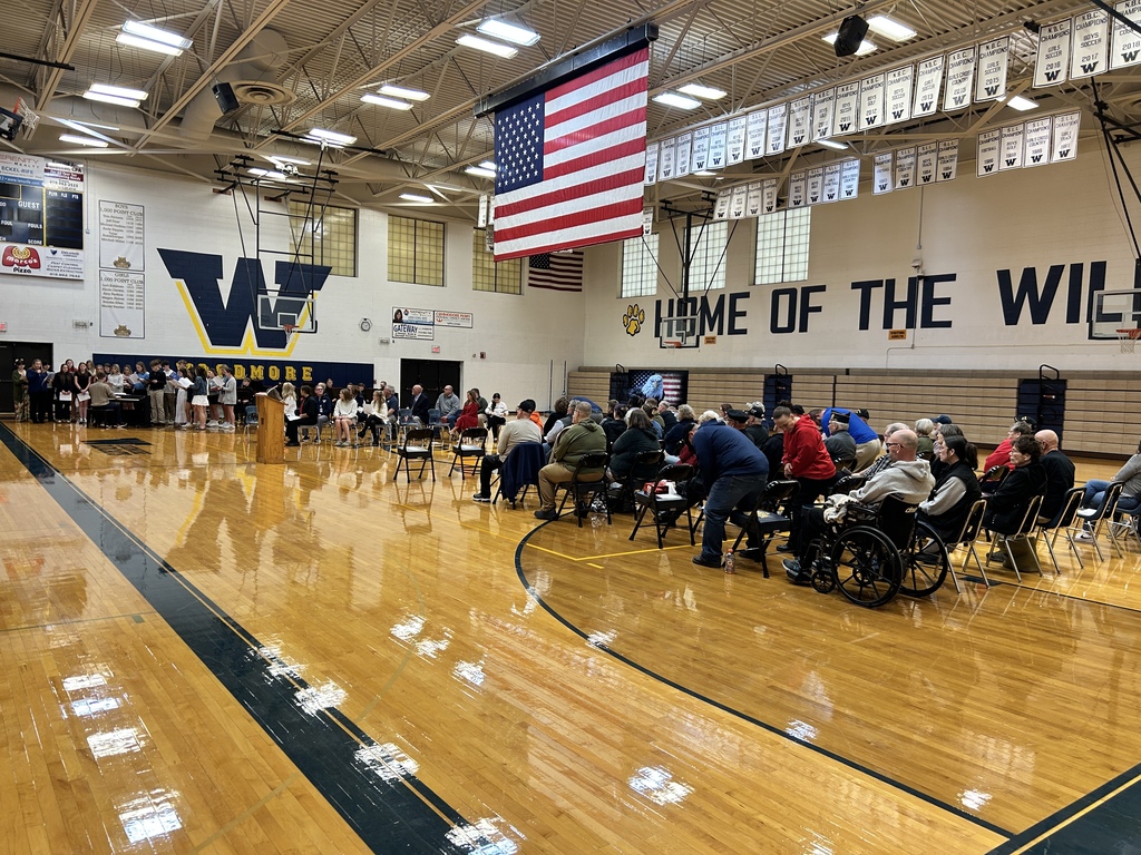 In the Woodmore gym, Veterans are seated on the right of the picture, with their backs to the camera, watching the Choir sing. The choir is at the other end of the gym, singing around a piano being played by their teacher, Mr. Fullenkamp. A large American flag is hanging from the ceiling in the middle of the gym.