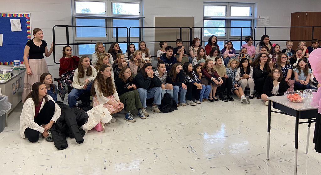 Sixth grade students sit on choir risers to listen to "Effie" (Mrs. Kistner) select the students names for the "Reaping."
