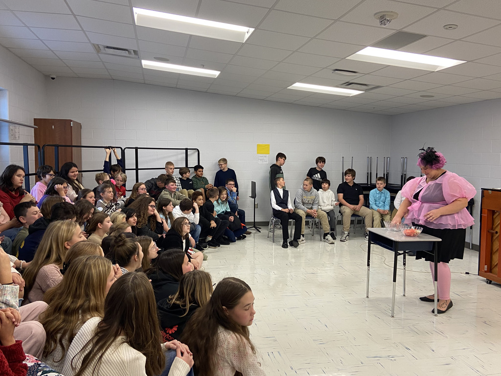 Sixth grade students sit on choir risers to listen to "Effie" (Mrs. Kistner) select the students names for the "Reaping."