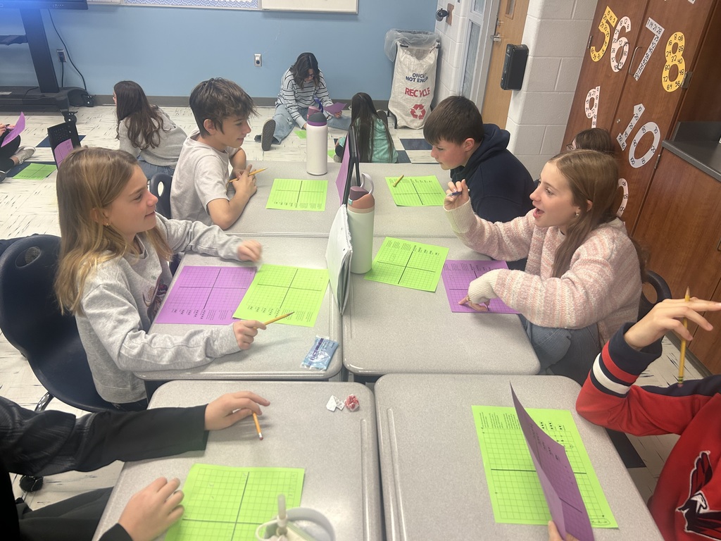 Four sixth grade students play coordinate grid battleship in pairs by sitting across from each other. Four more students sit on the floor in the background, also playing the game.