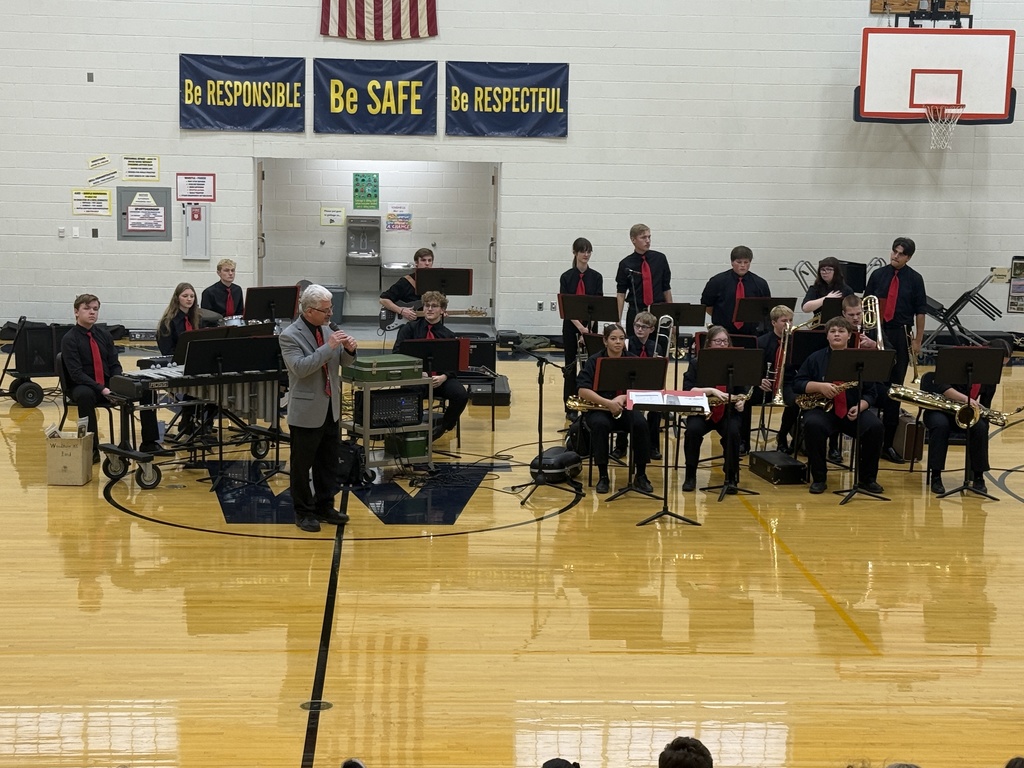 Our jazz band performs in the elementary/middle school gym. They are dressed in all black with red neck ties. Mr. Emerine, our band conductor, stands in front explaining the songs into the microphone.
