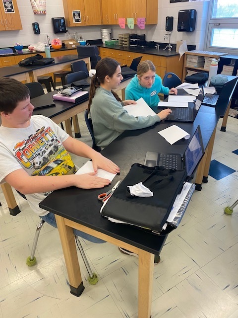 Three seventh grade students are back at their desk making revisions to their paper airplanes and looking up information on their Chromebooks.