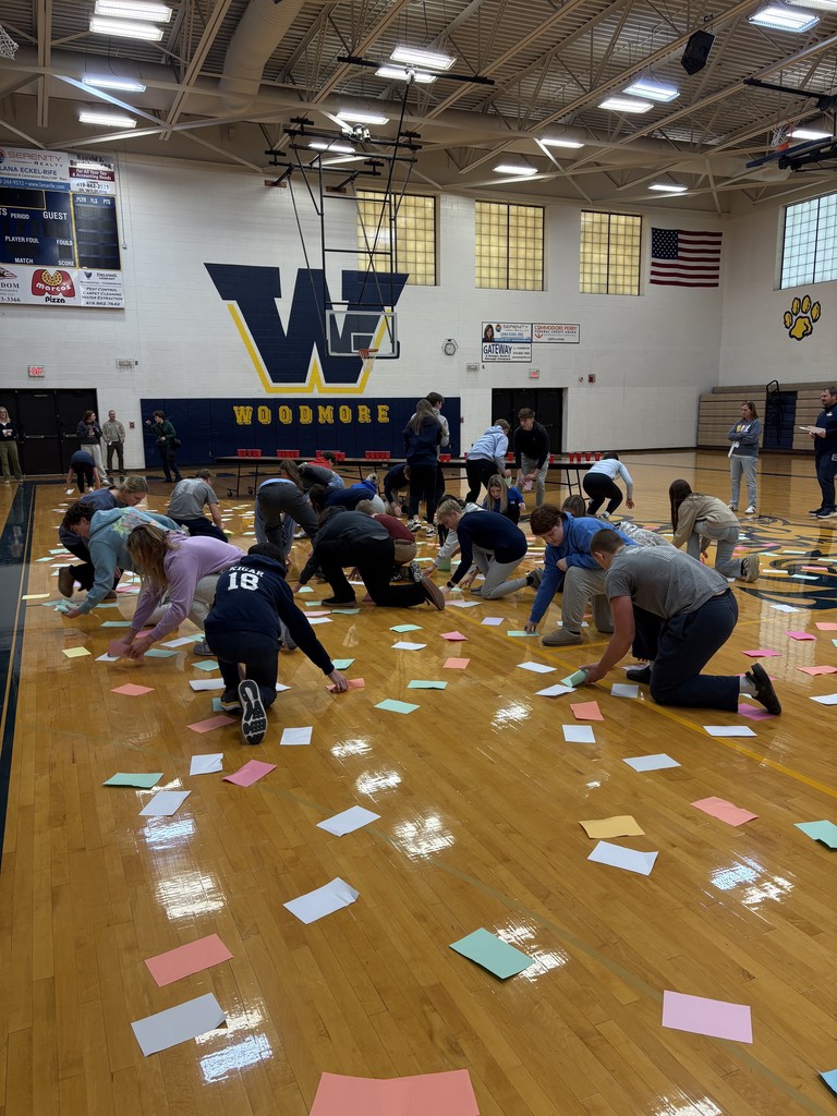 Students are on the gym floor looking at pieces of colored paper as part of their games. There are pieces of paper strewn everywhere!