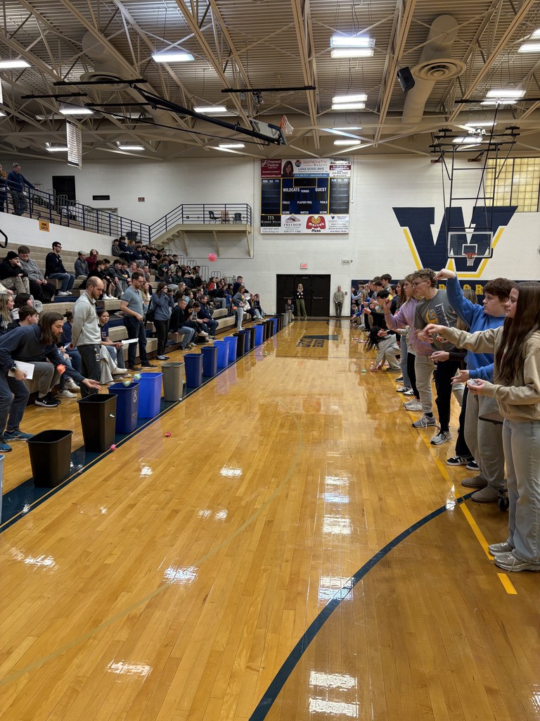 Participating students are lined up down the length of the gym floor all trying to toss an object into a trash can located some distance away with teachers watching to make sure the objects are making it in the cans. Other students sit in the bleachers watching the games.