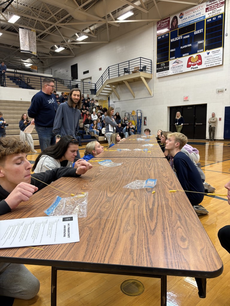 Students are kneeling in front of folding tables that are set up on the gym floor with their hands behind their backs. They have a piece of spaghetti in their mouth and they are attempting to pick up penne pasta from the table using only the piece of spaghetti. Teachers watch over as referees with students watching from the stands.