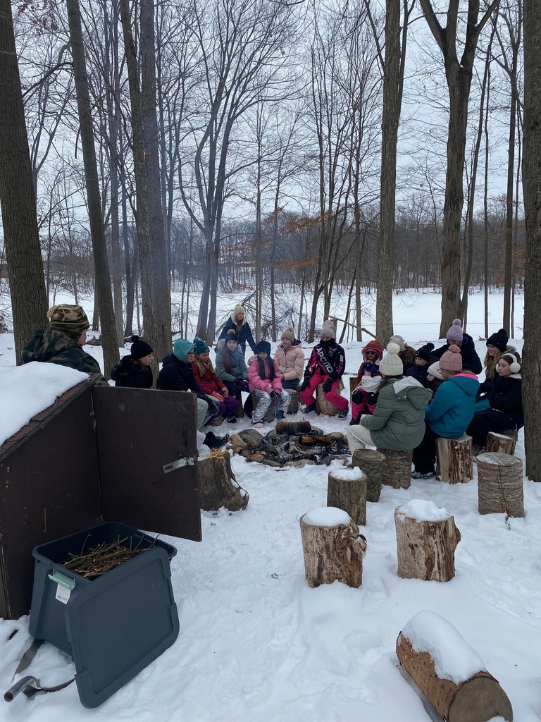 A groups of sixth grade students sit on tree stumps around a fire during an outdoor activity. There is quite a bit of snow on the ground!