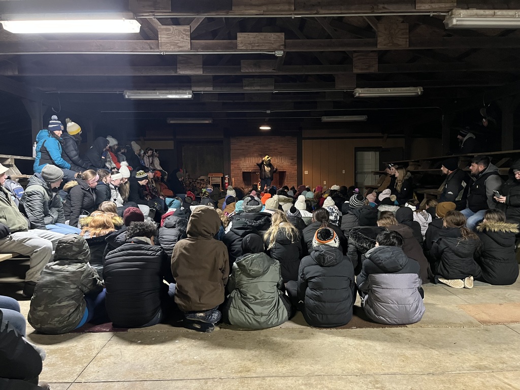 The sixth graders are all together outside for evening activities on their first night at camp. The area is lit with overhead lighting and appears to have a fireplace at the end where the presenter is standing and all of the students are looking.
