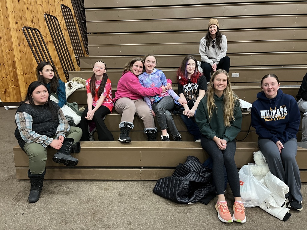 A group of sixth grade girls (plus two  high school cabin leaders and a teacher) sit on the bleachers inside after arriving to camp.