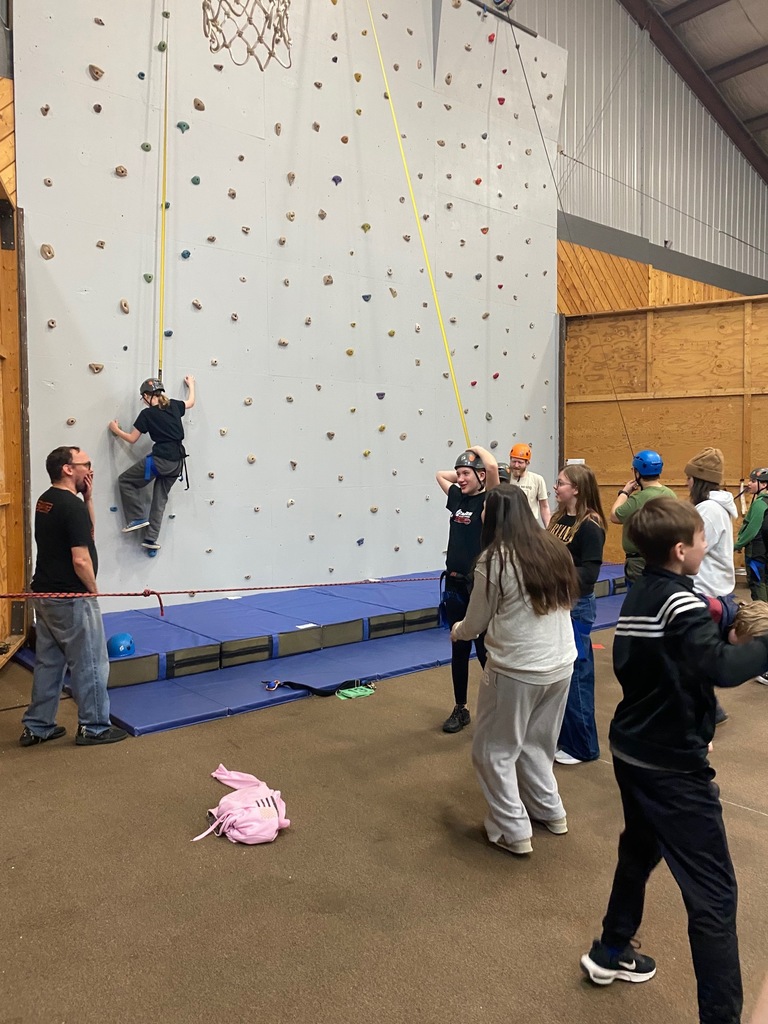 Students get geared up to climb on the climbing wall (one has already begun). There is a rope preventing them from accessing the wall until the instructors tell them they are ready.