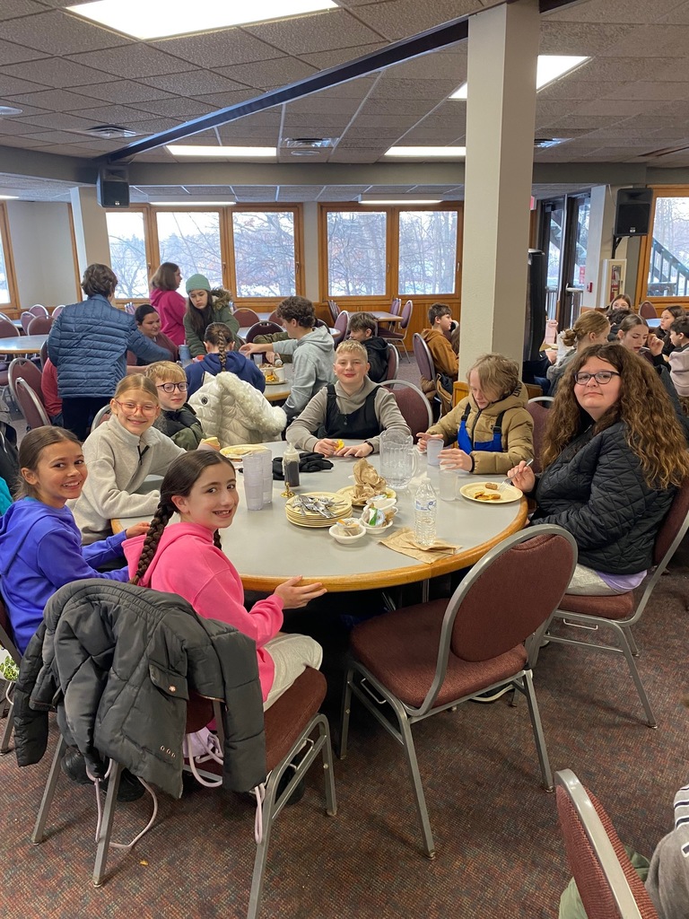 A group of sixth graders sitting around their table eating their meal. They're looking at the camera and posing.