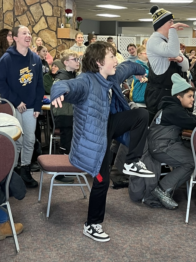 Students and cabin leaders are in the dining hall performing silly activities before their meal. There are several students standing and posing with general laughter from everyone in the picture.