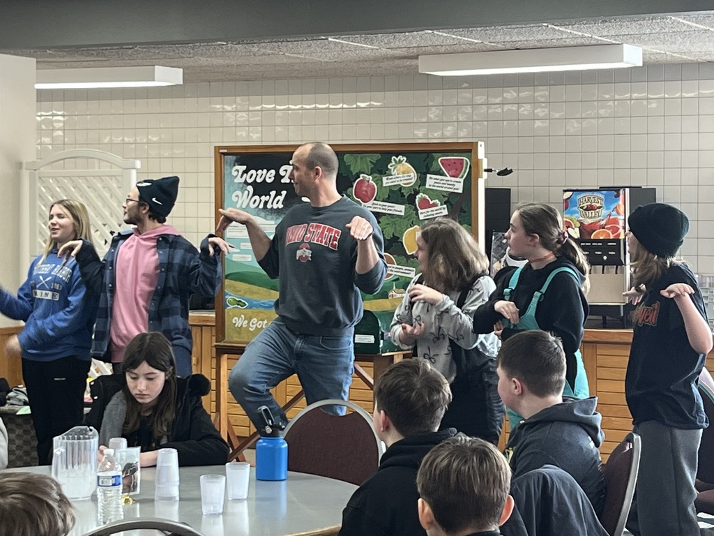 Sixth grade students (and Mr. Schlea and Mr. Kigar) are at the front of the dining hall participating in activities before their meal.
