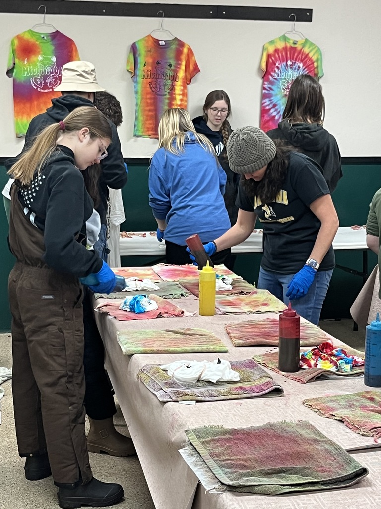 Students (and Mrs. Floro!) work on tie-dying shirts. There are multiple bottles of dye on the table. The shirts are balled up on the table and students are wearing protective gloves while completing the activity.