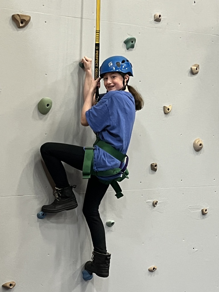 A sixth grade student turns around and smiles while on the climbing wall. She is wearing a helmet and harness.