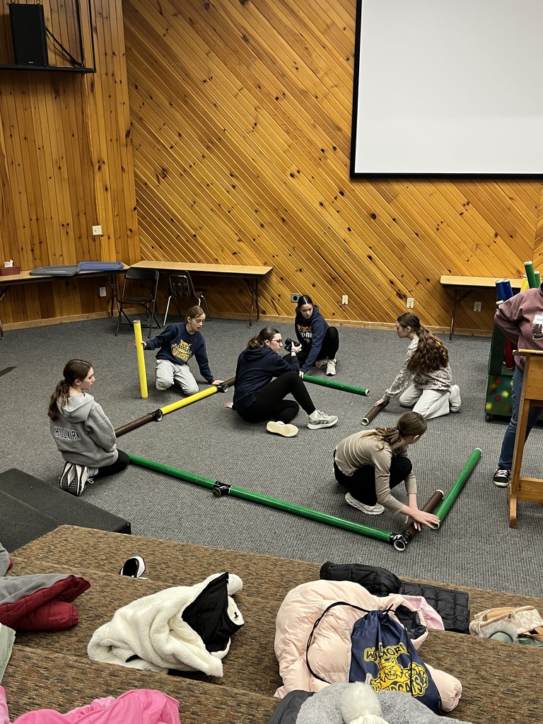 Six sixth grade girls work together on a puzzle challenge. They are inside, working on a carpeted floor, moving large colored tubes around and appearing to build something with them. 
