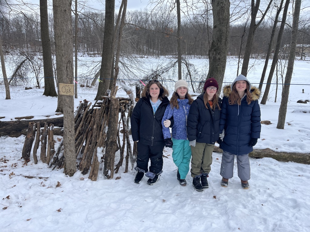 Four sixth grade girls pose in front of a shelter they built out of smaller tree limbs in the snow.