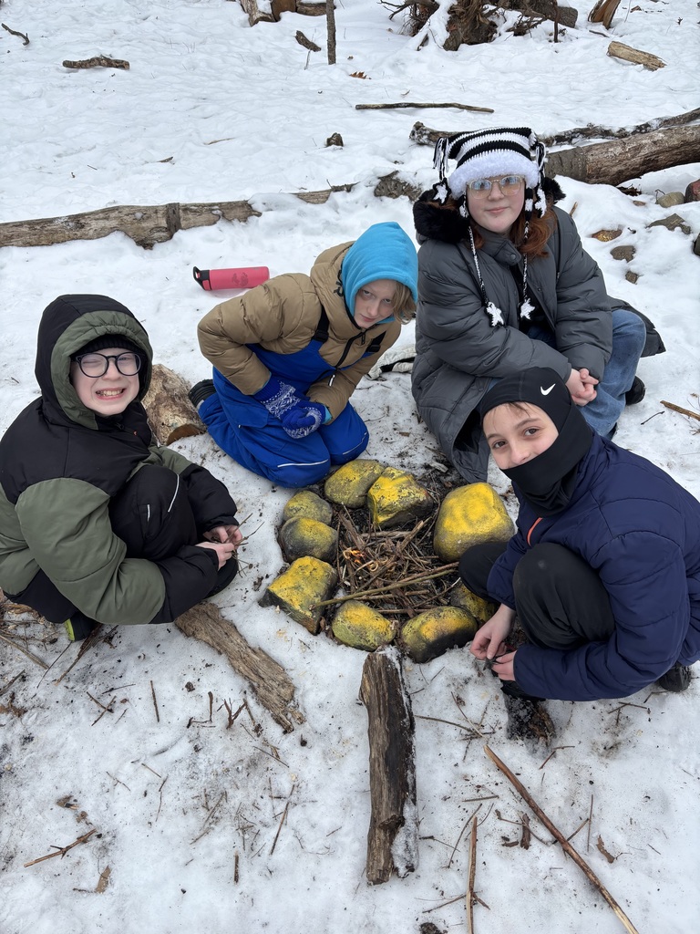 Four sixth grade students sit around the fire that they built. The fire is in a small pit that is surrounded by large rocks that have been painted yellow. There is still snow on the ground.