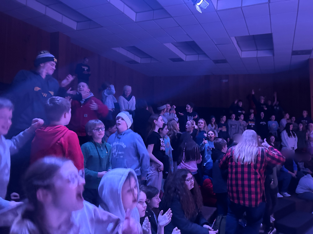 A group of sixth grade students and a few high school cabin leaders are dancing at a dance party inside. The lights are low and there is a purple/blue cast indicating that there might be dance lights somewhere in the room.